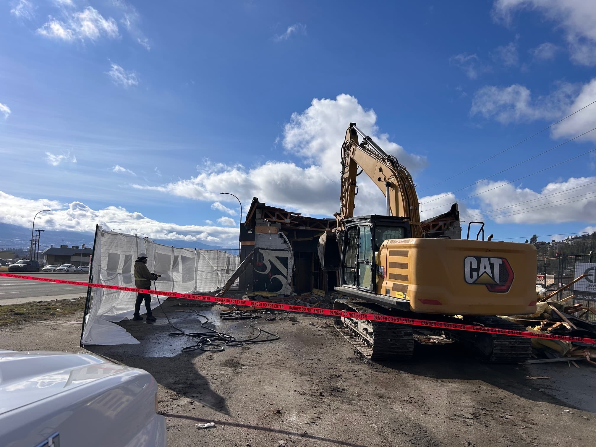 Yellow excavator at a demolition site with a worker spraying water behind red danger tape.